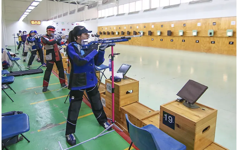 On the image, the frontal shot of an athlete pointing 
                            their rifle to a firing range its appreciated. The athlete 
                            is dressed in the discipline equipment. They are 
                            carrying a helmet, glasses, a vest and munitions.